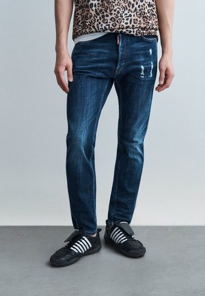 Személy sötétkék, karcsúsított farmernadrágot visel, enyhe kopott hatással a combnál, fekete cipőkkel, melyeken fehér csíkok láthatók.