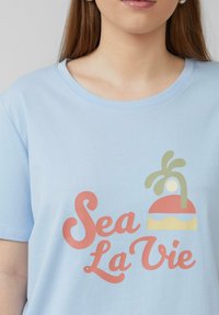 Lichtblauwe katoenen t-shirt met een grafisch ontwerp waarin "Sea La Vie" in roze schrift staat, vergezeld van een kleurrijke strandscene illustratie.