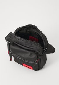 Borsa di nylon nera con due scomparti con zip, patch con logo rosso e tracolla regolabile. Texture liscia con un design minimalista.