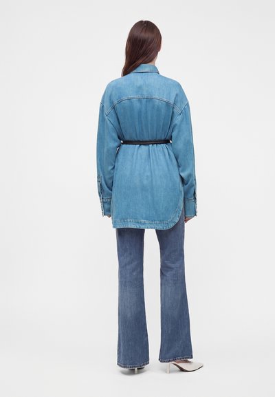 Femme portant une longue chemise en jean ceinturée à la taille, associée à un jean flare bleu et des chaussures blanches à talons, debout, vue de dos.