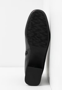 Chaussure en cuir noir vue par la semelle avec une semelle extérieure en caoutchouc texturé et un talon carré, reposant contre un coin blanc.