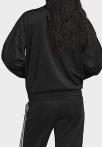 Svart polyesterrånarjacka med långa ärmar, elastiska cuffar och nederkant, tillsammans med svarta byxor med tre vita ränder på sidorna.