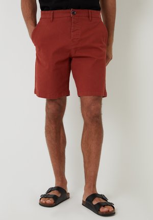 Shorts en coton rouge avec une fermeture à bouton, des poches latérales et une coupe droite, associés à des sandales noires avec deux sangles et une boucle.
