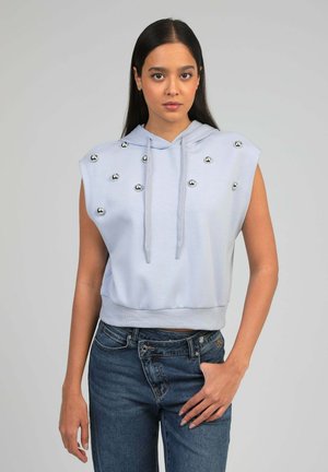 Femme portant un sweat à capuche sans manches bleu clair avec des clous métalliques, associée à un jean bleu, debout sur un fond gris uni.