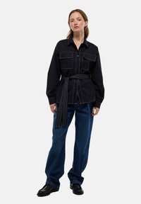 Chemise noire à boutons avec deux poches avant, couture contrastante et ceinture nouée. Associée à un jean en denim ample et des chaussures noires.