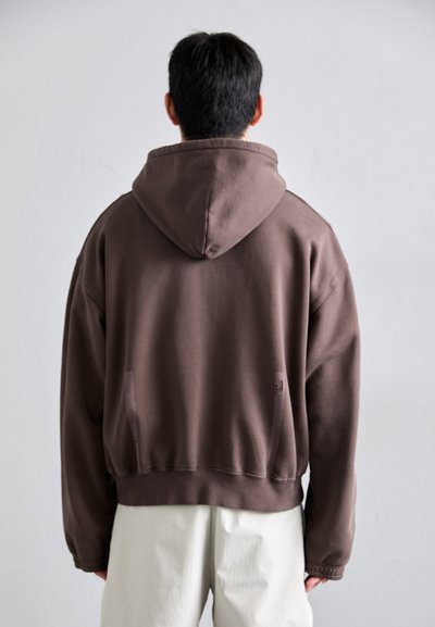 Sudadera con capucha marrón de corte holgado, con puños acanalados y bolsillos. Textura suave y color sólido, mostrada desde la parte posterior sobre un fondo neutro.
