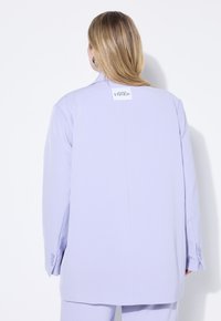 Blazer de gran tamaño en púrpura claro con solapa con muesca, una abertura trasera y acentos de botón a juego. Presenta una etiqueta de marca en la parte posterior.