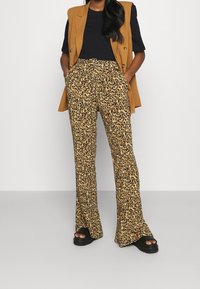 Helflairade byxor med leopardmönster i beige och svart, med hög midja och fickor, kombinerat med en svart skjorta och en brun väst.