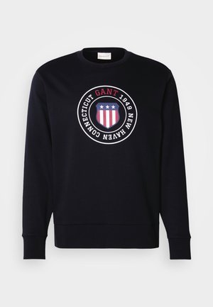Tummansininen college-paita, jossa on pyöreä logo, joka sisältää kilven, kolme tähteä, punaiset ja valkoiset raidat sekä teksti "GANT 1949 New Haven Connecticut."