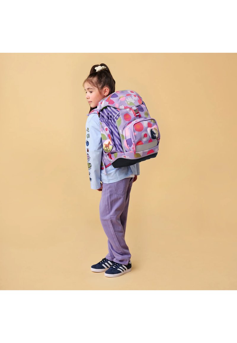 Junges Mädchen in violetten Hosen und hellblauem Hemd trägt einen großen bunten Rucksack mit Tierapplikationen auf beigem Hintergrund.