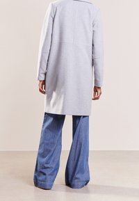 Světlý šedý dlouhý vlněný kabát s přizpůsobeným střihem a kapsami s rozparkem, ve spojení s širokými džínovými kalhotami. Hladká textura a čisté linie.