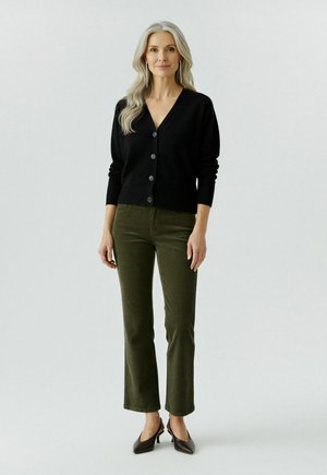 Femme aux longs cheveux blonds portant un cardigan noir à boutons, un pantalon vert olive et des talons aiguille noirs, se tenant devant un fond uni.