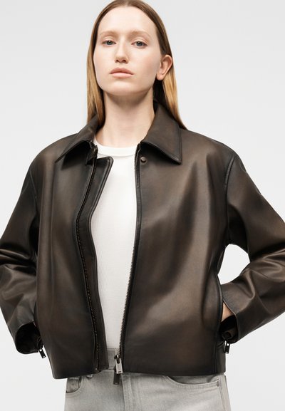 Γυναίκα που φοράει μία σκούρα καφέ δερμάτινη ζακέτα πάνω από λευκό μπλουζάκι και ανοιχτόχρωμα γκρι τζιν, στέκεται με τα χέρια στις τσέπες μπροστά σε ένα μονόχρωμο φόντο.