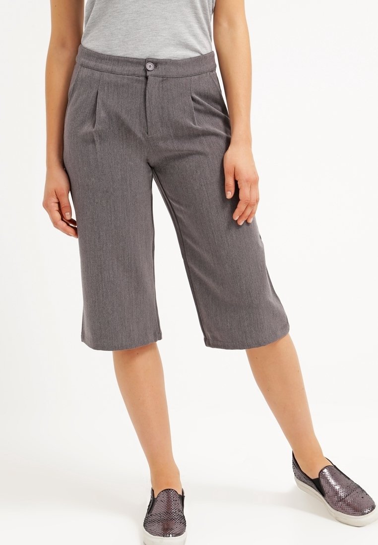 Femme portant un short gris sur mesure mi-longueur avec des plis et une taille boutonnée, assorti à des chaussures argentées sans lacets.