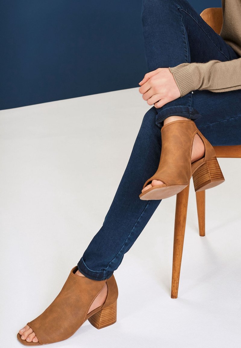 Sandales à talons ouvertes marron avec un design à bout ouvert et un talon en bois texturé, associées à un jean slim bleu foncé. Matière en cuir souple.