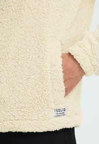 Pullover in pile color crema con una superficie morbida e intrecciata. Presenta una tasca laterale e un'etichetta all'orlo.
