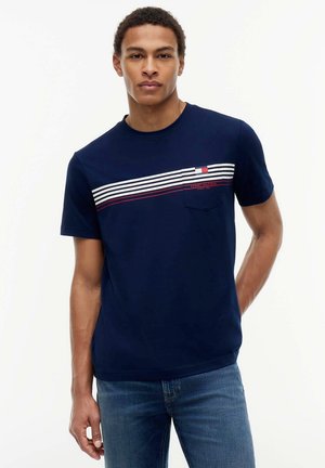 Jonge man draagt een marineblauw T-shirt met witte en rode horizontale strepen over de borst en blauwe spijkerbroek, staand tegen een effen achtergrond.