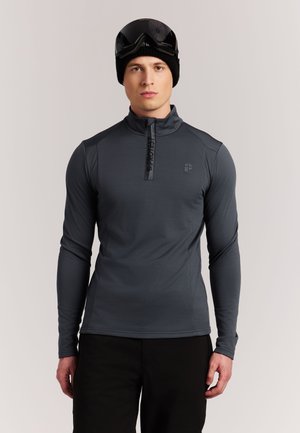 Jonge man die een donkergrijze longsleeve met halve rits, zwarte broek, zwarte muts en skibril op zijn voorhoofd draagt, kijkt vooruit.