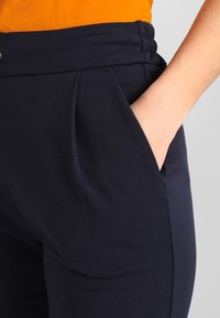 Mörkblå byxor gjorda av mjukt tyg, med hög midja, sidofickor och subtila veck för extra designdetalj.