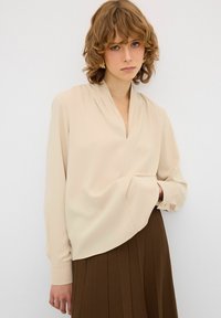 Beige Bluse mit langen Ärmeln, V-Ausschnitt und plissiertem Design. Weiches Material mit glatter Textur, das einen leicht asymmetrischen Saum aufweist.