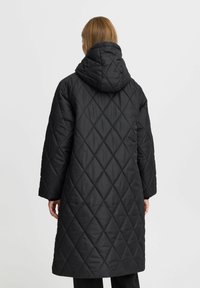 Manteau long matelassé noir avec capuche, présentant des motifs en losange, des manches larges et une texture lisse. La vue arrière montre aucun détail supplémentaire.