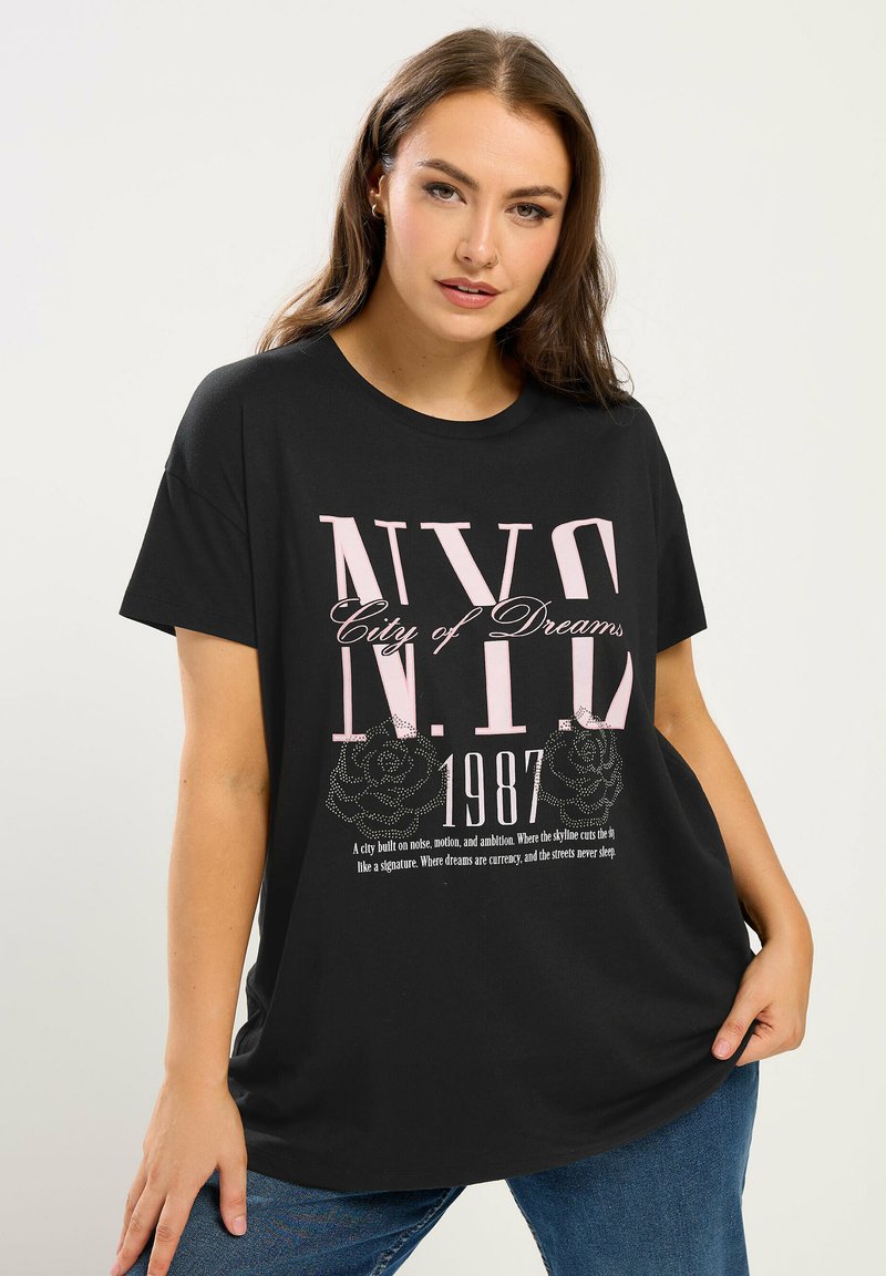 Junge Frau trägt ein schwarzes, übergroßes T-Shirt mit pinker Aufschrift "NYC City of Dreams 1987" und Rosengrafiken, kombiniert mit blauer Jeans.