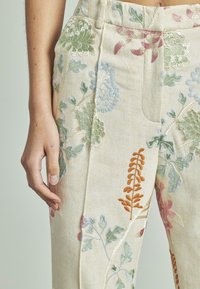 Lato sinistro di una persona che indossa pantaloni color crema con ricami floreali multicolori, mano rilassata ai lati, contro uno sfondo semplice.