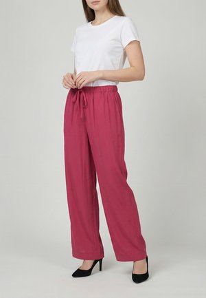 Mujer vestida con una camisa blanca de manga corta, pantalones rosas sueltos con cordón y zapatos negros de tacón alto, de pie frente a un fondo liso.