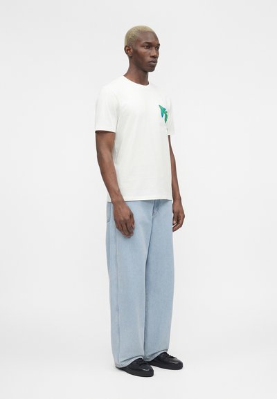 Άνδρας με κοντά ξανθά μαλλιά που φοράει λευκό μπλουζάκι με πράσινο σχέδιο πεταλούδας και ανοιχτό μπλε φαρδιά τζιν, στέκεται πλαγίως.