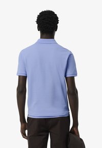 Sinih polo paita dengan kerah klasik dan lengan pendek, terbuat dari kain halus, dipakai dengan celana gelap, dilihat dari belakang.