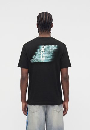 Άτομο με κοτσίδες που φορά μαύρο μπλουζάκι με λογότυπο «Oakley» με εφέ glitch στην πλάτη και ανοιχτόγκρι τζιν.