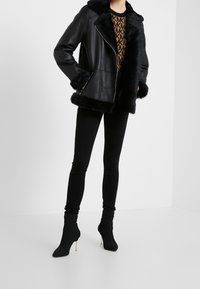 Chaqueta de cuero negra con ribete de piel sintética, cierre de cremallera y grandes solapas. Usada sobre una blusa marrón con estampado, combinada con leggings negros y botas de tobillo.
