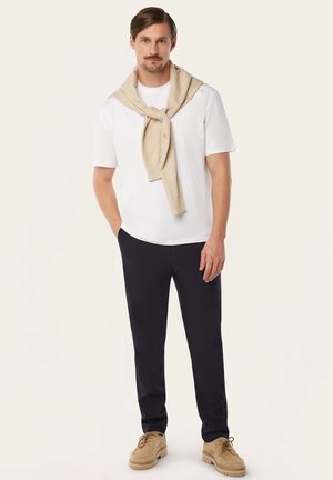 Mann trägt weißes T-Shirt, beigen Pullover um die Schultern gebunden, schwarze Hose und beige Schuhe, steht mit einer Hand in der Tasche vor einfachem Hintergrund.
