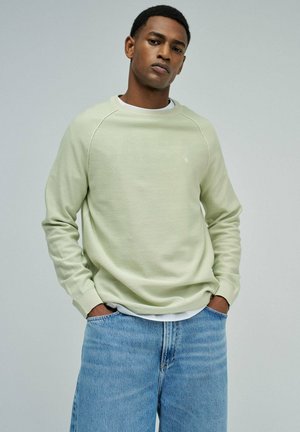 Joven con cabello corto, vistiendo un suéter verde claro y pantalones vaqueros azules, de pie con las manos en los bolsillos contra un fondo liso.