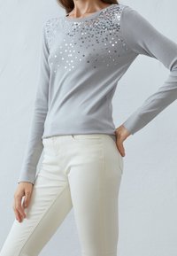 Maglietta a maniche lunghe grigia chiara con paillettes argentate brillanti sul petto. Tessuto morbido, design aderente, scollatura tonda e polsini a costine.