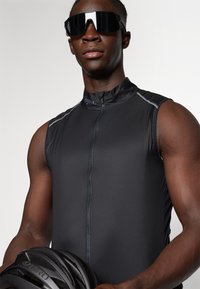 Maillot de cyclisme noir sans manches avec une fermeture éclair à l'avant et des coutures réfléchissantes. Le modèle porte des lunettes de soleil noires surdimensionnées et tient un casque noir.
