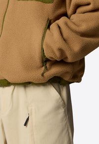 Giacca in pile beige con dettagli verdi, tasche con zip e polsini elasticizzati, abbinata a pantaloni color crema con tasca laterale con zip.