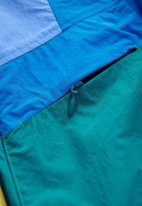Gros plan sur une veste en tissu bleu et turquoise avec une fermeture éclair noire visible et une tirette de fermeture sur une poche.