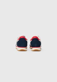 O pereche de adidași navy blue, albaștri și roșii, văzuți din spate, cu tălpi de culoare gumă, pe un fundal alb simplu.