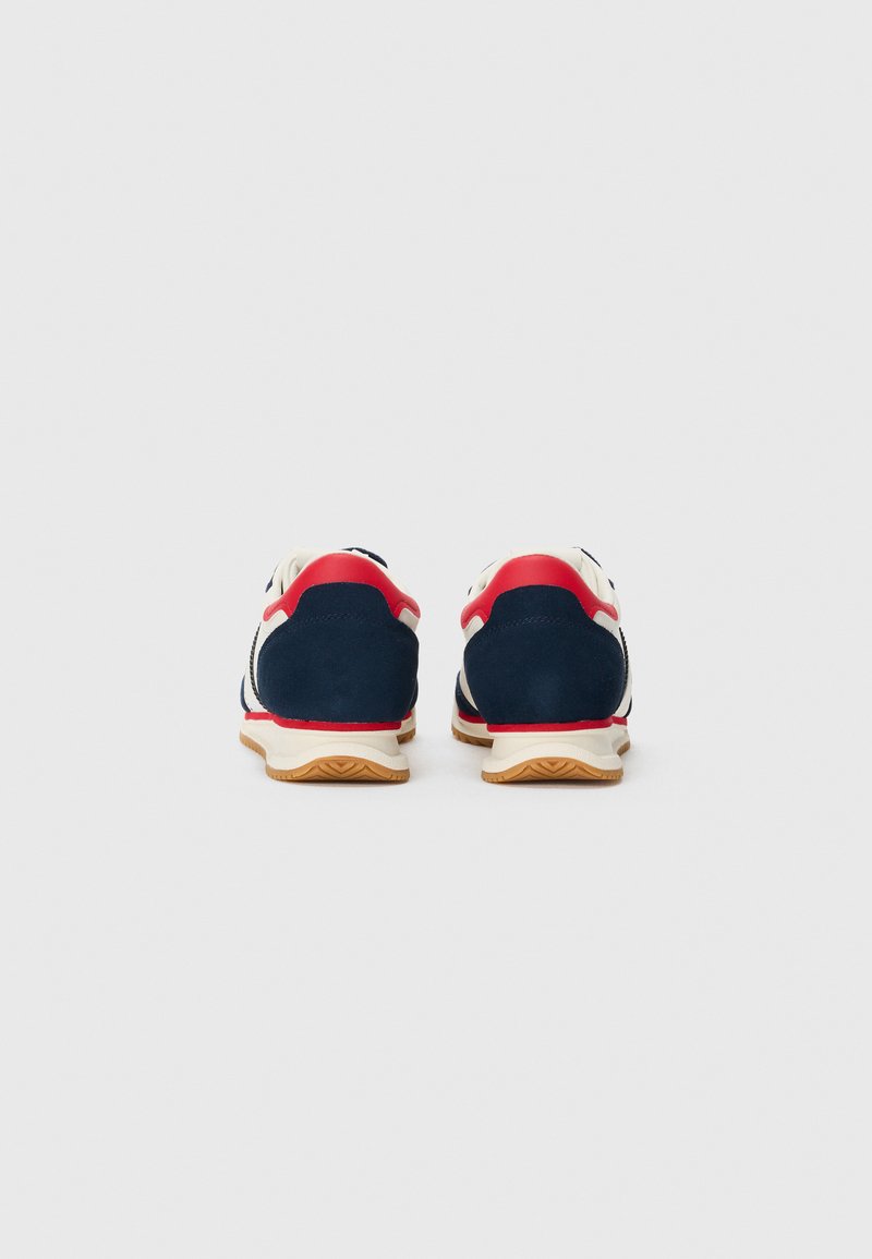 Une paire de chaussures de sport bleu marine, blanches et rouges vues de l'arrière avec des semelles couleur gomme sur un fond blanc uni.