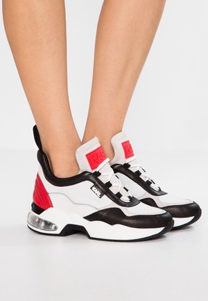 Osoba stojící na bílé ploše, obuvena v bílých, černých a červených robustních teniskách s viditelnými podrážkami s air cushion, s nápisem „Karl“ na bocích a jazycích.