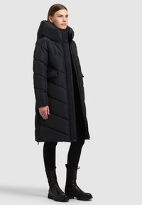 Must pufferjope kõrge kraega, millel on diagonaalsed tikitud mustrid, kaks esitaskut ja lühis lukuga. Modell kannab musti pahkluu saapaid.