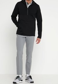 Pull noir à capuche avec un zip mi-long, associé à un pantalon gris clair et des baskets noir et blanc avec des accents turquoise.