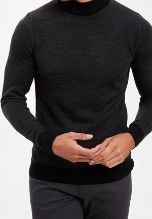 Uomo che indossa un maglione dolcevita grigio scuro con tessuto strutturato e bordo nero, mani delicatamente intrecciate in vita, su uno sfondo semplice.