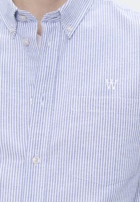 Light blue and white striped button-up shirt, featuring a chest pocket and a small white logo on the pocket area. Soft fabric texture.