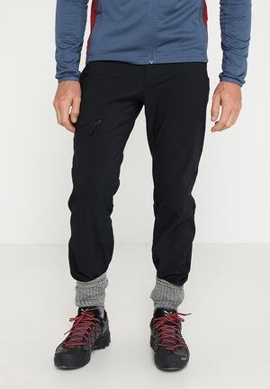 Uomo che indossa pantaloni neri con cerniera, calzini grigi in maglia, scarpe da trekking nere con lacci rossi e una giacca blu con pannelli laterali rossi.