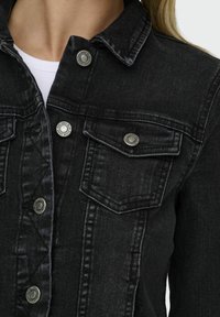 Svart denimjacka med en klassisk design, silverfärgade knappar, två bröstfickor och en texturerad yta med synlig sömnad.