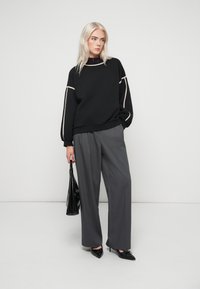 Ξανθιά γυναίκα στέκεται μπροστά σε λευκό τοίχο φορώντας μαύρο πουλόβερ με μπεζ ρίγες, φαρδιά γκρίζα παντελόνια, μαύρες γόβες και κρατώντας μαύρη τσάντα.