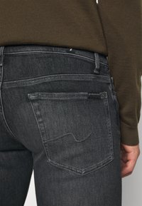 Primer plano de una persona que lleva vaqueros gris oscuro con un bolsillo trasero cosido y una camiseta de manga larga marrón metida por dentro.