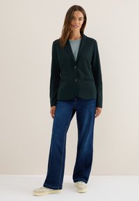 Donkergroene blazer met twee knopen, notch revers en voorzakken, gecombineerd met een lichtblauwe gebreide top en blauwe wijde spijkerbroek.